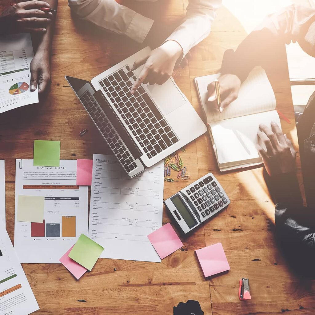 A team of professionals viewed from above, working at a table covered in printed charts, calculators, sticky notes, and a laptop.